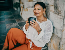 A woman sits on a tile floor, holding a mug as she leans against a stone interior wall.