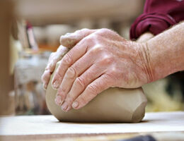 A pair of hands molding clay on a work surface with a jar of paintbrushes in the background.