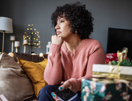 A woman with a melancholy expression sits on a couch. Holiday lights and gifts are nearby.